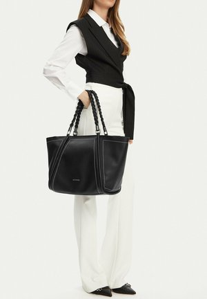 Kobieta w białych spodniach i koszuli z czarną kamizelką trzyma dużą czarną skórzaną torbę typu tote z plecionymi uchwytami.