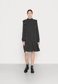 Vestido preto com bolinhas, com mangas longas, ombros com volantes e bainha em camadas. Usado com botas pretas até o tornozelo, apresentando detalhes em metal circular.