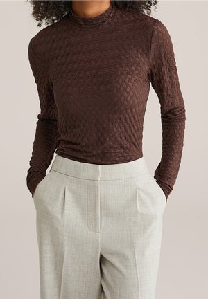 Personne portant un haut marron texturé ajusté à manches longues, rentré dans un pantalon plissé beige clair taille haute, les mains dans les poches.