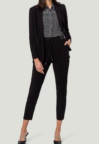Blazer noir sur mesure porté par-dessus un chemisier noir et blanc à motifs, associé à un pantalon noir ajusté et des chaussures à talons avec des accents en chaîne.