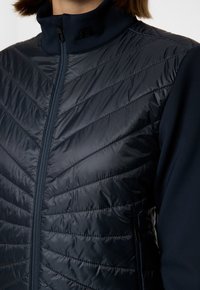 Marinblå jacka med kviltat mönster, med hög krage, dragkedjeslutning och texturerade paneler. Inkluderar sidofickor och en logotypdetalj.