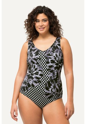 Smilende kvinde med krøllet hår iført en sort og hvidternet badedragt med store lilla blomster på en lys baggrund.