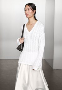 White knitted sweater with a V-neck, featuring vertical ribbed texture. Worn with a flowing white skirt and a small black bag.