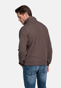 Bruine sweatshirt met textuur en hoge kraag, voorzien van een subtiel patroon en een logo-opdruk op de linkermouw. Gekocht in combinatie met blauwe spijkerbroek.