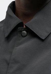 Črna jakna s teksturirano tkanino in ovratnikom. Na jakni je črn gumb z izbočenim logotipom. Prikaz bližnjega posnetka ovratnika in detajla gumba.