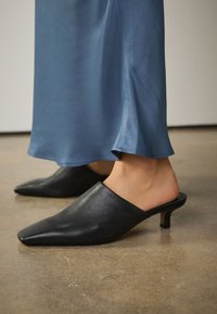 Mules en cuir noir à bout pointu et talon kitten, portées avec une jupe longue en satin bleu jusqu’à la cheville sur un sol lisse.