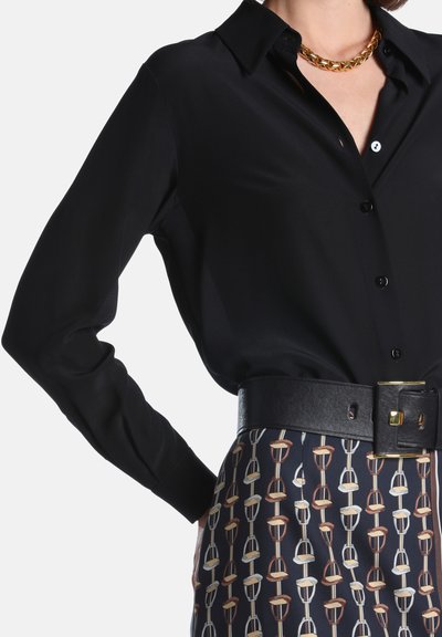 Femme portant un chemisier noir boutonné, un collier chaîne dorée épais et une jupe à motifs avec une large ceinture noire.