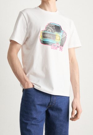 Person trägt ein weißes T-Shirt mit buntem Vintage-Auto-Motiv und dem Text "HUGO est. '93", kombiniert mit blauen Jeans, Hand in der Tasche.