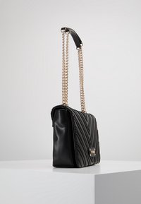 Czarna skórzana torba na ramię z pikowanym wzorem w zygzak, złotym łańcuszkiem oraz prostokątnym złotym akcentem na froncie.