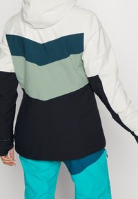 Venkovní bunda s kapucí; design s blokováním barev v bílé, tmavě modré, tyrkysové a zelené; obsahuje vzor v podobě šipek a elegantní siluetu.