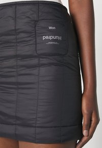Mini-jupe matelassée noire avec poche avant affichant le texte "Won Hundred" et les coordonnées, portée par une personne à la peau foncée et au bras détendu.