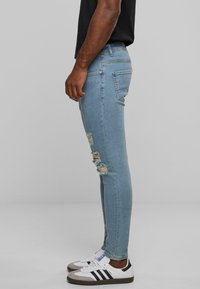 Vékony, világoskék farmernadrág, térdnél megfosztott szakadásokká, denim anyagból készült, fehér és fekete sportcipővel kombinálva, oldalról nézve.