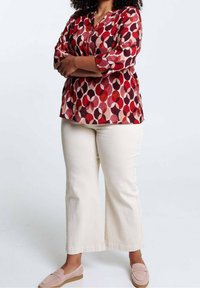 Donna con le braccia incrociate che indossa una maglietta a maniche lunghe rossa con motivo, pantaloni larghi color crema e scarpe slip-on rosa, in piedi su uno sfondo bianco.