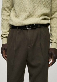 Pull en tricot texturé beige associé à un pantalon marron foncé et une ceinture noire. Le pantalon présente un pli et des poches latérales.