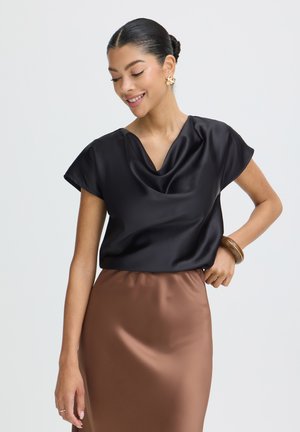 Naeratav naine, kes kannab musta satiinsärki lühikeste varrukatega, pruuni seelikut, kuldseid kõrvarõngaid ja pruuni käevõru, taustaks on lihtne taust.