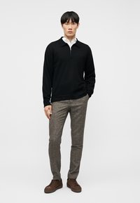 Άνδρας που φοράει μαύρο πουλόβερ με φερμουάρ πάνω από λευκό πουκάμισο, γκρι στενά υφασμάτινα παντελόνια και καφέ αθλητικά παπούτσια, στέκεται μπροστά σε λευκό φόντο.