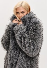 Grå faux pelsfrakke med høj krave, lange ærmer og tekstureret, fluffy materiale; har et lagdelt design og blød, tæt udseende.