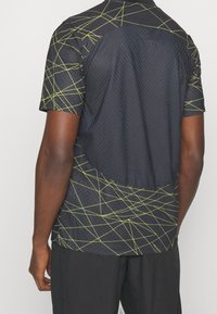 Maglietta nera a maniche corte con tessuto strutturato, caratterizzata da un motivo geometrico di linee gialle e un design con apertura rotonda sulla schiena.