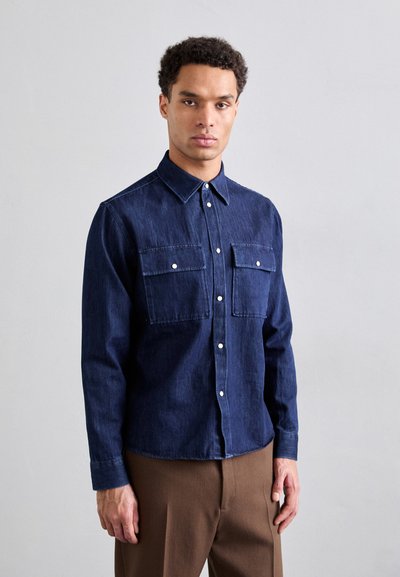 Cămașă din denim albastru închis, cu două buzunare pe piept, guler tip buton și închidere cu nasturi, asortată cu pantaloni maro. Textură netedă.