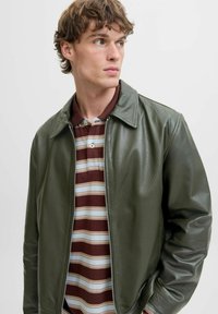 Chaqueta de cuero verde con cremallera frontal, cuello y puños acanalados, superpuesta a una camiseta polo marrón con rayas. Textura suave y diseño ajustado.