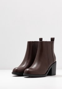 Brązowe skórzane botki za kostkę z żebrowanym elastycznym panelem, okrągłym noskiem i grubym obcasem. Gładka faktura i kontrastowa czarna podeszwa.