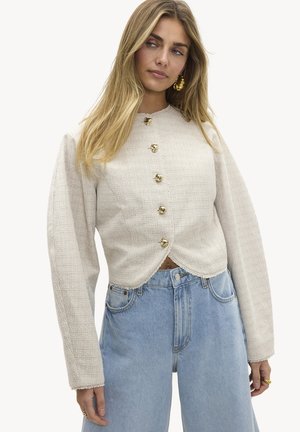 Vrouw met lang blond haar, gekleed in een crèmekleurig, gestructureerd jack met gouden knopen en lichtblauwe jeans met een hoge taille.