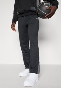 Harmaat urheiluhousut, joissa on valkoiset sivuraitat, valmistettu sileästä kankaasta. Yhdistettynä valkoisiin tennareihin, pitävät mustaa koripalloa.