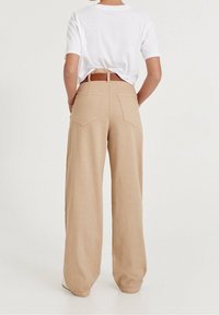 Person wearing beige wide-leg pants with back pockets, brown belt, white t-shirt tucked in, standing against a plain background.