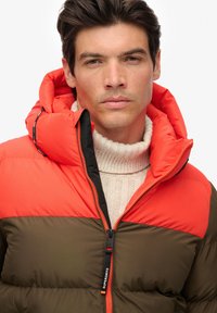 Puffer jacket with an orange hood and shoulder panels, brown body, and a black zipper. Features a textured knit sweater underneath.