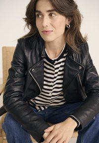 Veste en cuir noir avec fermeture éclair argentée, portée sur une chemise à rayures noir et blanc. Jean en denim bleu visible. Environnement à texture douce.
