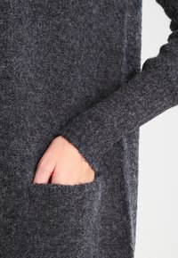 Šedý vlněný svetr s texturovaným povrchem, dlouhými rukávy a přední kapsou, s detailním záběrem ruky odpočívající v kapse.