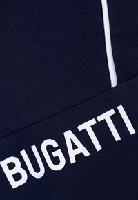Tessuto blu navy con la scritta "BUGATTI" in grandi lettere bianche. Il tessuto è liscio con una leggera elasticità, rifinito da una striscia di accento bianca.