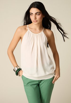 Kvinde med langt mørkt hår iført hvid ærmeløs top, grønne bukser og chunky armbånd, poserer mod en beige baggrund.