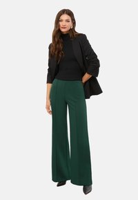 Zaļas platajās bikses ar priekšējiem šuvumiem, apvienotas ar pieguļošu melnu ribotu tērpu un melnu žaketi. Melnas augstpapēžu kurpes noslēdz izskatu.