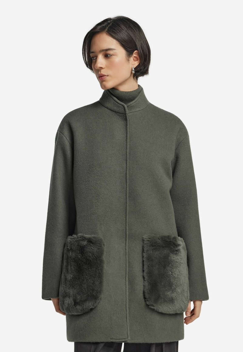Överdimensionerad olivgrön kappa i ullblandning med hög krage, med två stora fickor i konstfuskpäls, slät textur och minimal knäppning.