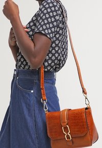 Bolso bandolera de cuero con grabado de cocodrilo en color marrón, de forma estructurada, con herrajes dorados y una correa ajustable, combinado con una blusa estampada.