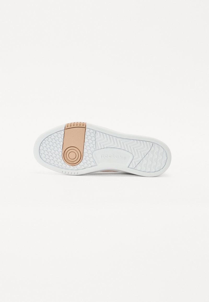 Suela de una zapatilla con una base de goma blanca, patrón de tracción en espiga, y un acento marrón con logo en relieve en el talón.