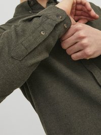 Camisa de franela verde oliva con cuello y puños abotonados, que presenta una tela texturizada y botones marrones, mostrando el brazo en movimiento.
