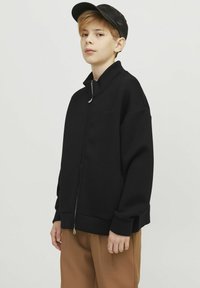 Melns zip-up jaka ar augstu apkakli, ribotām piedurknēm un gludu tekstūru, kombinēta ar brūnām biksēm un melnu cepuri.