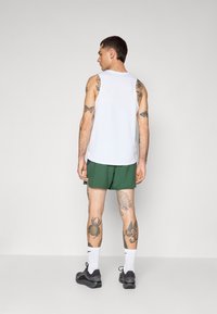 Top sin mangas blanco, shorts verdes, zapatillas negras y calcetines blancos. Tatuajes visibles en brazos y piernas. Fondo liso.