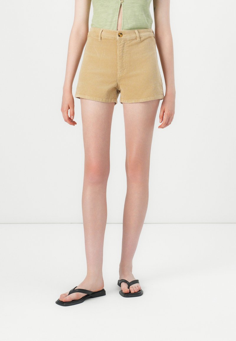 Beige corduroy shorts with a high waist, front button closure, and two side pockets. Paired with black flip-flops. Smooth texture.