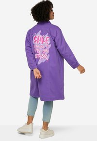 Paarse lange jas van katoen met lange mouwen, voorzien van roze graffiti-achtige tekst op de rug, gestyled met lichtblauwe jeans en witte sneakers.