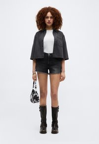 Νεαρή γυναίκα με κατσαρά μαλλιά φοράει μαύρο κοντό σακάκι, άσπρο τοπ, μαύρες τζιν σορτς, μαύρες μπότες και κρατά τσάντα με ασπρόμαυρο μοτίβο.