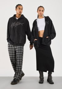 Two people with braided hair wear black zip-up hoodies; one pairs with plaid pants and boots, the other with a white crop top, black skirt, and boots.