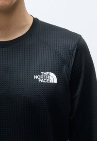 Schwarzes Sportshirt mit langenÄrmeln, das ein strukturiertes, perforiertes Muster und ein weißes Logo auf der linken Brust aufweist. Rundhalsausschnitt und Raglanärmel.