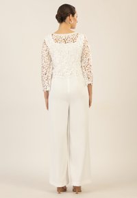 Weißer Jumpsuit mit einem Spitzenoberteil, das mit floralen Mustern und dreiviertellangen Ärmeln verziert ist. Weit geschnittene, ausgestellte Hosen runden das Design ab.