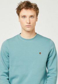 Giovane uomo con capelli castano chiaro mossi che indossa un felpa azzurro chiaro con un piccolo logo di un albero marrone sul petto, rivolto verso l'avanti su uno sfondo neutro.