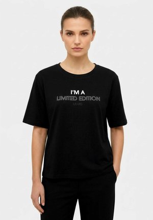 Donna con i capelli legati indossa una maglietta nera con scritta bianca "I'M A LIMITED EDITION" su uno sfondo chiaro e uniforme.