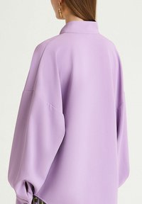 Blusa oversize lilla con colletto, maniche ampie e leggero arricciatura sui polsini. Tessuto liscio e vestibilità morbida.