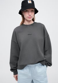 Sudadera de color gris oscuro con cuello redondo y logotipo bordado, combinada con pantalones cortos de textura azul claro. El modelo lleva un sombrero tipo cubo negro.
